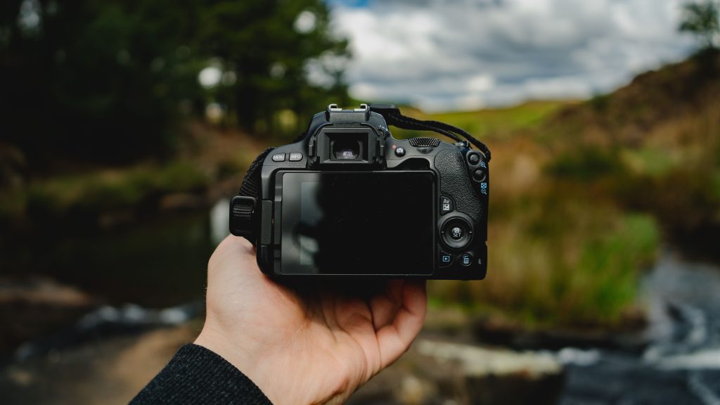 Close-up of a hand holding a camera in an outdoor setting with nature in the background.